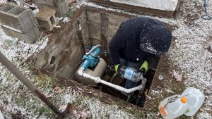 In the center is a square hole in the ground, about chest deep.  The top dimensions are the same.  It's surrounded by concret blocks and tools.  In the hole is a water pump and some pipes. A person strands in the hole pouring water into a pipe.  It's snowing.