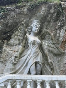 A stone relief sculpture of an angel carved into a rocky cliff face. The angel has large, detailed wings and flowing hair, wearing a draped garment that gives a sense of movement. Her arms are extended slightly outward with open hands, and her head is tilted gently downward. Moss and weathering mark parts of the sculpture, and a white balustrade runs across the bottom foreground. Green plants grow along the top edge of the rock above.