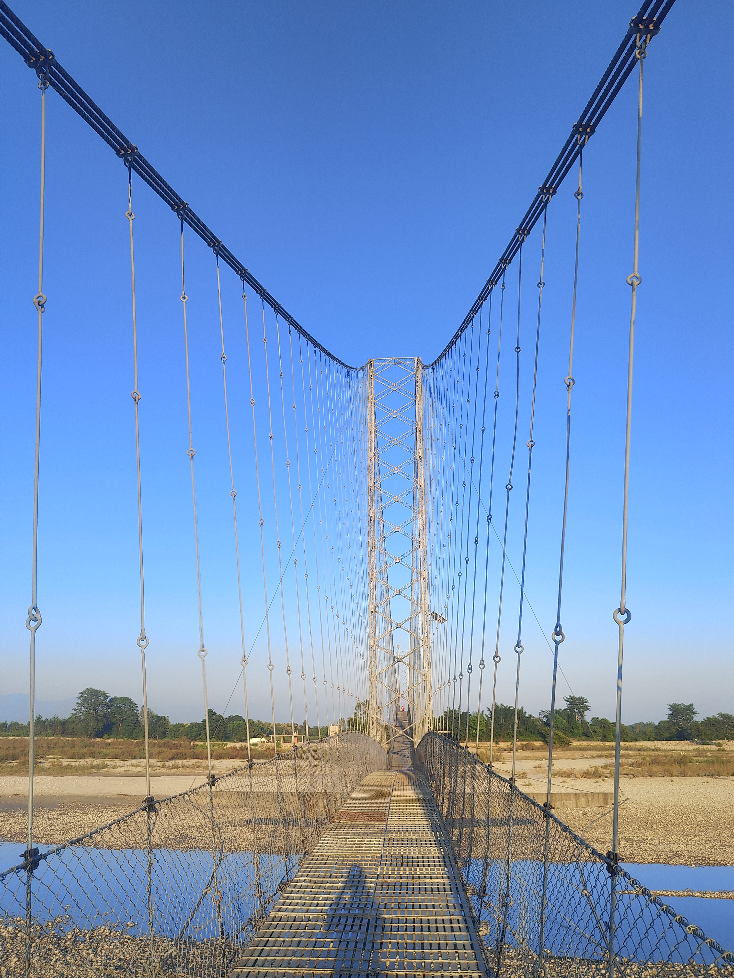 A suspension bridge stretches across a dry riverbed under a clear blue sky.
