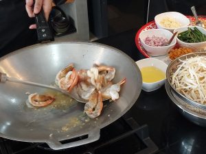 The shrimp being stir-fried in a wok, with Pad Thai ingredients like bean sprouts, eggs, and chopped vegetables prepared on the side.