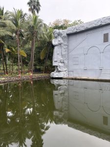 A gray stone statue of Swami Vivekananda beside a seated figure, reflected in a dark green pool, surrounded by tall palm trees.