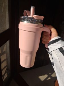 This image shows a person holding a large, pastel pink insulated tumbler with a handle, a clear lid, and a straw, all illuminated by warm sunlight coming from the side.