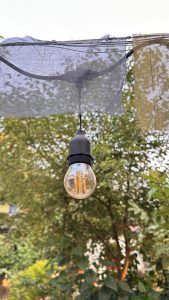 A vintage-style light bulb hangs from a wire, surrounded by soft fabric drapes in shades of gray and yellow. In the background, a lush green garden is visible, adding a serene and natural ambiance to the scene.
