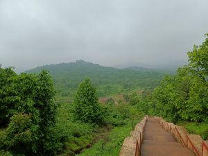 A stone pathway descending through dense green forest, leading toward mist-covered hills under an overcast sky, captures a calm and scenic natural landscape.