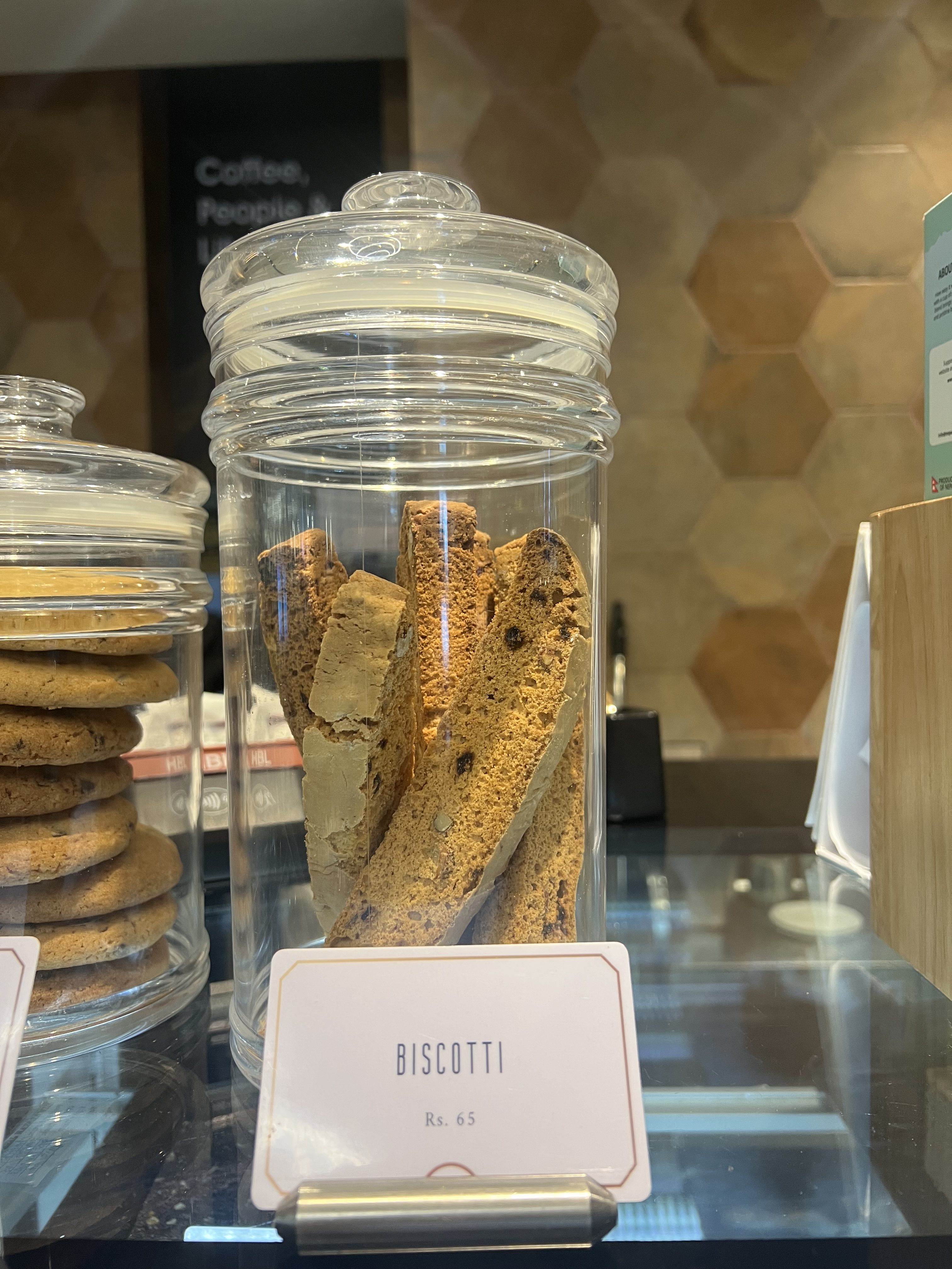A tall, clear glass jar on a counter holds several long pieces of biscotti, with a small sign in front labeling it "BISCOTTI Rs. 65