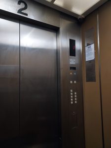 The inside of an elevator, showing metal doors, a control panel with floor buttons, and a digital floor display. The space looks compact and modern, with neutral-colored walls.
