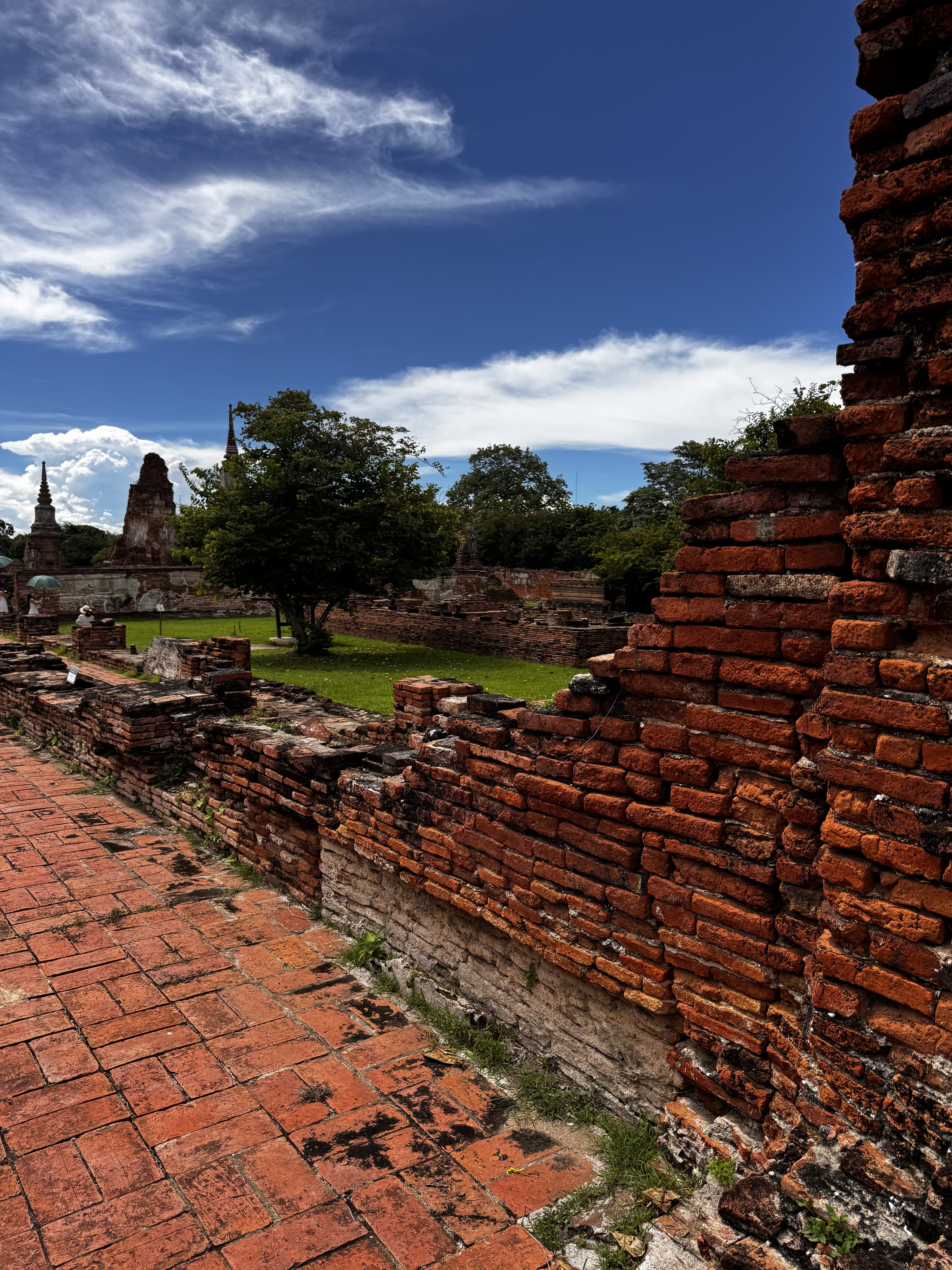 A partially collapsed brick wall with weathered red bricks is in the foreground, leading to a grassy area surrounded by ruins in the background. There are scattered trees and remnants of brick structures, with a bright blue sky dotted with fluffy white clouds above.