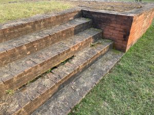A series of weathered brick steps leading up to a raised platform, surrounded by grass.