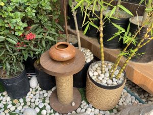 A small wooden table with a round top holds a traditional clay pot. Surrounding the table are various plants, including green leaves and small red flowers, as well as taller bamboo stalks in a black pot filled with smooth pebbles. 