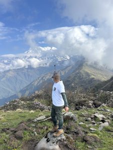 A person stands on a rock in a mountainous landscape, looking out at a view of snow-capped peaks under a partly cloudy sky