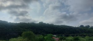 A landscape view of dense green hills under a cloudy sky, with layers of trees covering the hillside and a small red-roofed building visible at the bottom. Soft sunlight filters through the clouds, creating a peaceful natural scene.