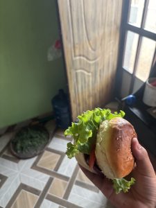 A hand holding a hamburger with lettuce and tomato, set against a background featuring a partially open wooden door and a tiled floor.