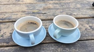 Two light blue cups of coffee sit on small matching saucers, placed on a weathered wooden table. 