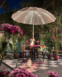 This photograph captures an intimate, carefully staged outdoor dining scene set at night, designed to evoke romance, calm, and warmth. At the center is a small round table for two, placed beneath a large cream-colored patio umbrella. The umbrella is adorned with delicate string lights that radiate softly from the central pole, creating a gentle glo
