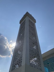 Tall minaret with intricate latticework reaches skyward under a bright sun. Clouds and a blue sky frame the tower, creating a serene atmosphere.