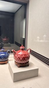 A decorative red teapot with a stylised design is displayed on a pedestal in a museum setting. 