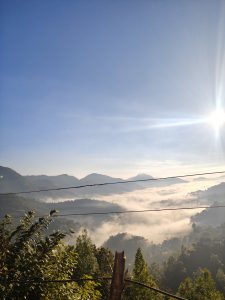 A scenic view of a mountain landscape shrouded in morning fog, with rolling hills and peaks in the background under a clear blue sky. 