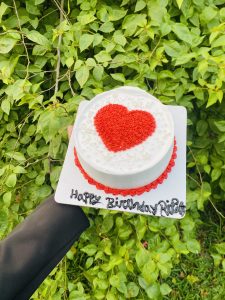 A hand holds a white birthday cake with a red heart, set against lush green leaves.