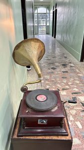 A vintage gramophone with a large brass horn is placed against a light green wall in a corridor. 
