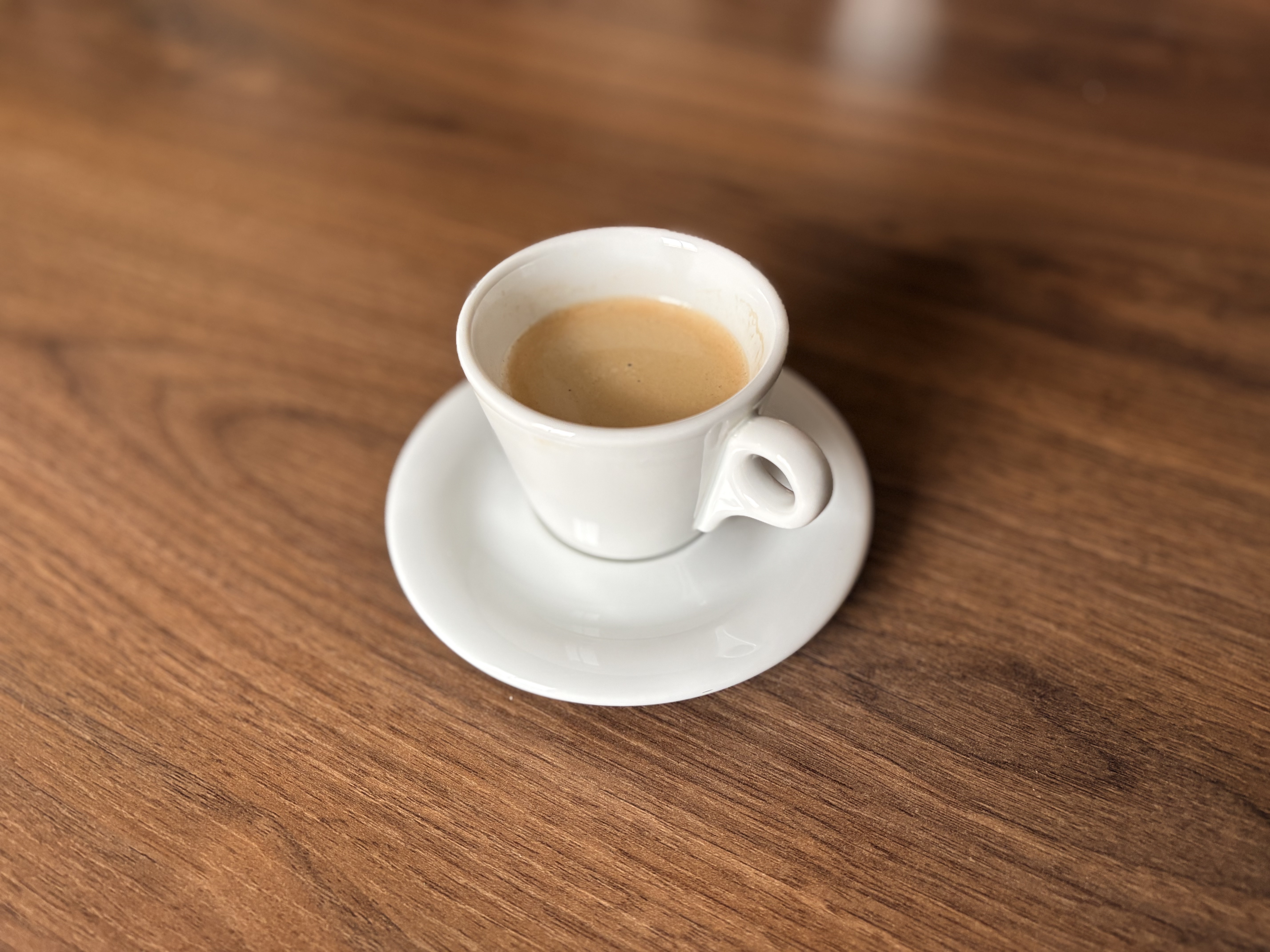 A small white ceramic espresso cup filled with coffee sits on a matching saucer, placed on a wooden table. The photo is taken from above at a slight angle, showing the coffee’s light crema. The background is softly blurred, emphasizing the cup and saucer.