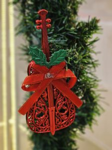 A decorative red violin-shaped ornament with a ribbon, hanging on a Christmas tree. Shot in Kozhikode, Kerala, the image captures festive spirit, holiday d&eacute;cor, and fine details with a pleasing background blur and vibrant seasonal colors. 