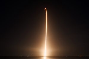 An orange light trail streaks into the dark night sky as a Falcon 9 rocket launches a commercial satellite. 