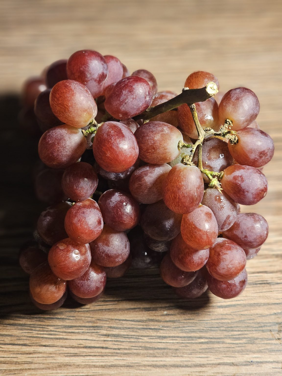 A cluster of fresh red grapes is placed on a wooden surface, showing soft light, natural shine, and rich color. The close-up view brings out the fruit’s texture and freshness in a warm and straightforward setting. Captured from Perumanna, Kozhikode.