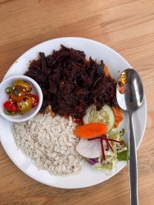 A white plate on a wooden table holds a serving of deep red shredded meat, a pile of white flattened rice (chiura), and a small salad with sliced vegetables, alongside a dish of chilies.