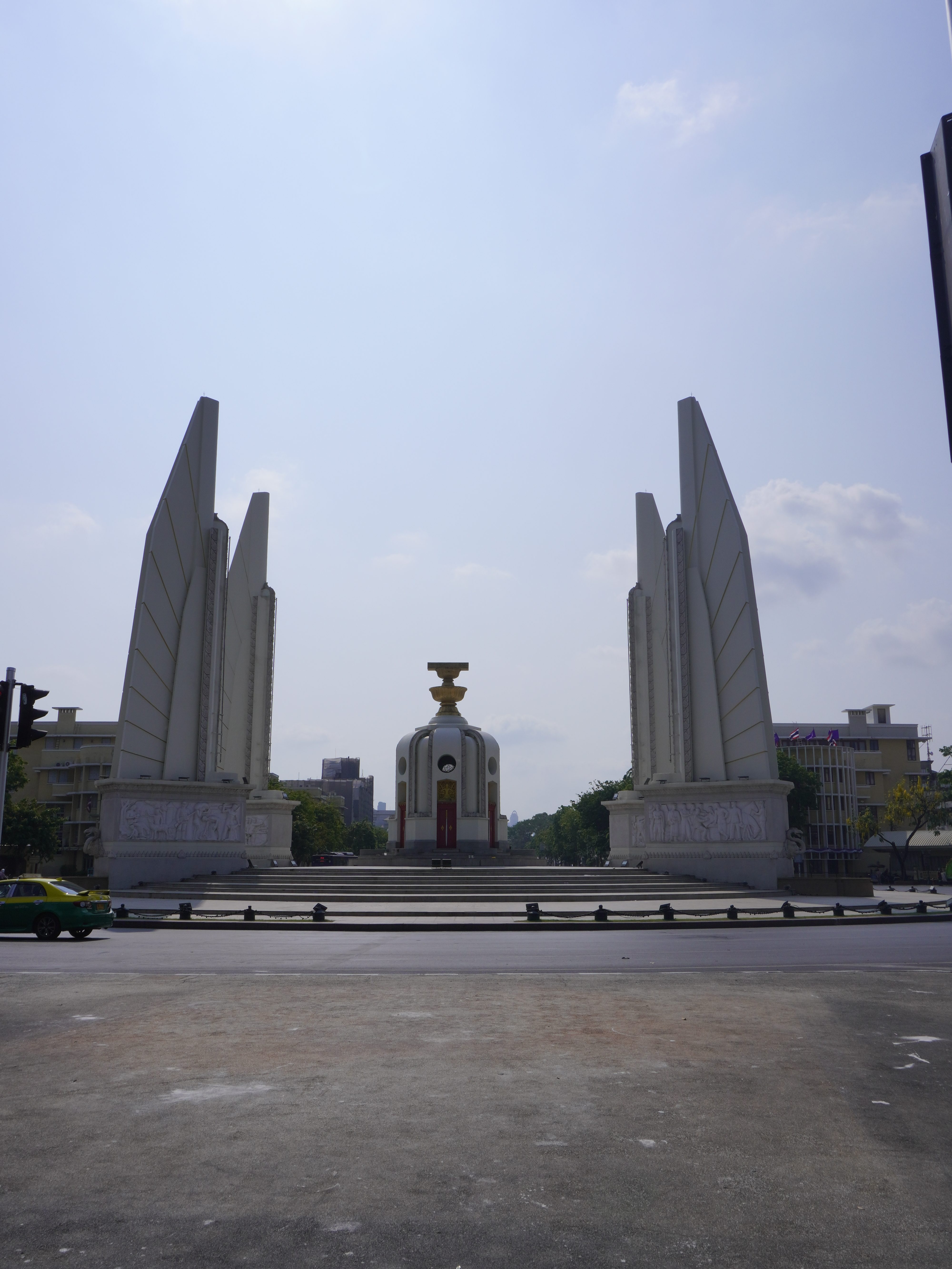 Photo of the Democracy Monument in Bangkok, Thailand. It is located on a roundabout on the Ratchadamnoen road. The center of the monument is manuscript of the constitution, surrounded by four pillars. The road before the monument is mostly empty.