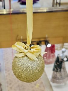 A glittering golden Christmas ornament with a soft yellow ribbon hangs indoors, reflecting festive lights. The simple decoration adds warmth and celebration to the season, photographed in Kozhikode, Kerala.