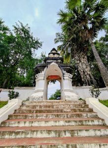 A frontal view of the ancient entrance arch of Vat Visounnarath in Luang Prabang, with weathered stone steps, tall palm trees, and lush greenery.

