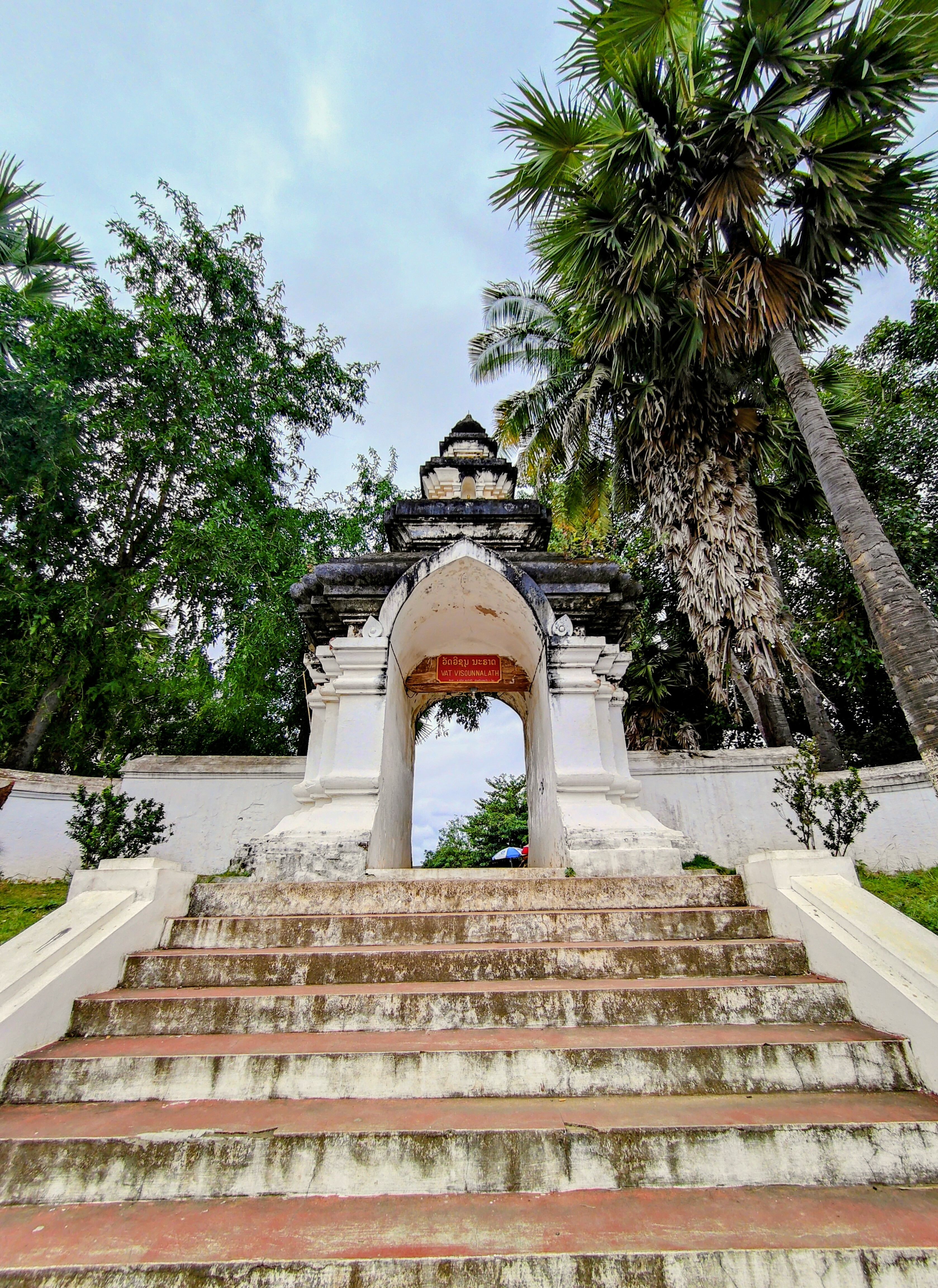 A frontal view of the ancient entrance arch of Vat Visounnarath in Luang Prabang, with weathered stone steps, tall palm trees, and lush greenery.