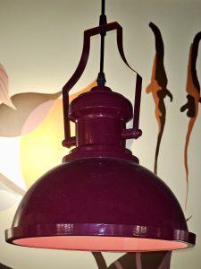 A close-up view of a vintage-style hanging lamp painted in deep maroon, glowing warmly against an artistic wall. Taken in Kozhikode, Kerala, the photo highlights indoor décor, soft light, and creative design details with a modern café vibe. 