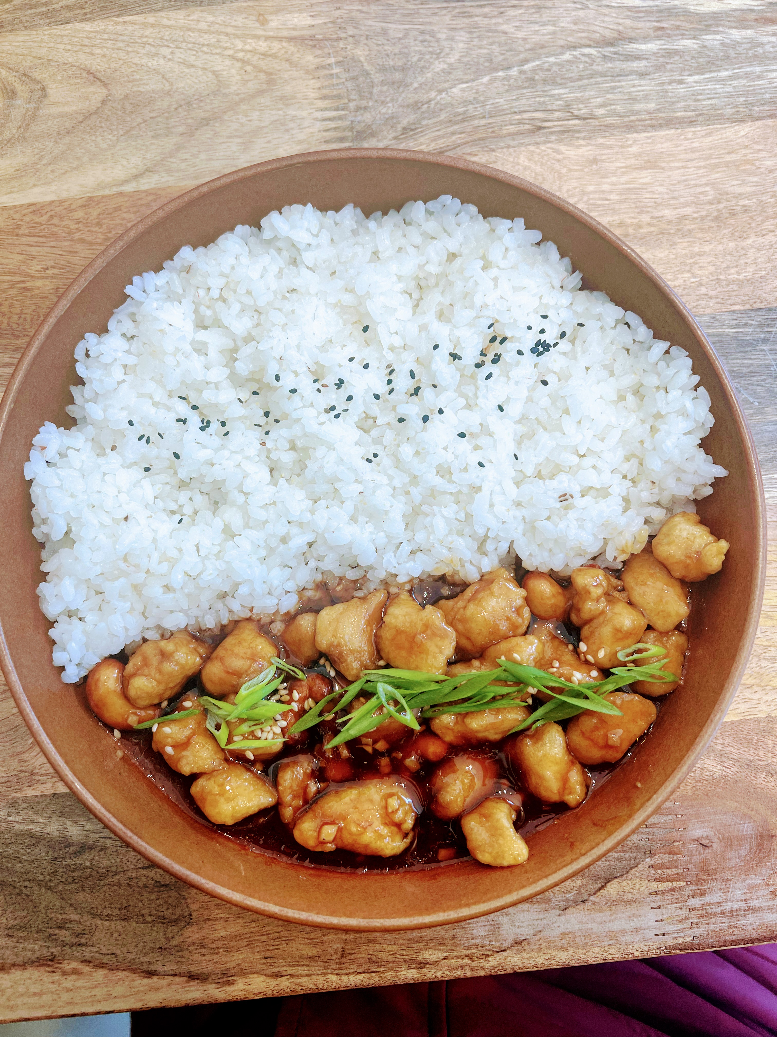 A bowl of white rice served with crispy chicken in sauce, topped with green onions and sesame seeds.