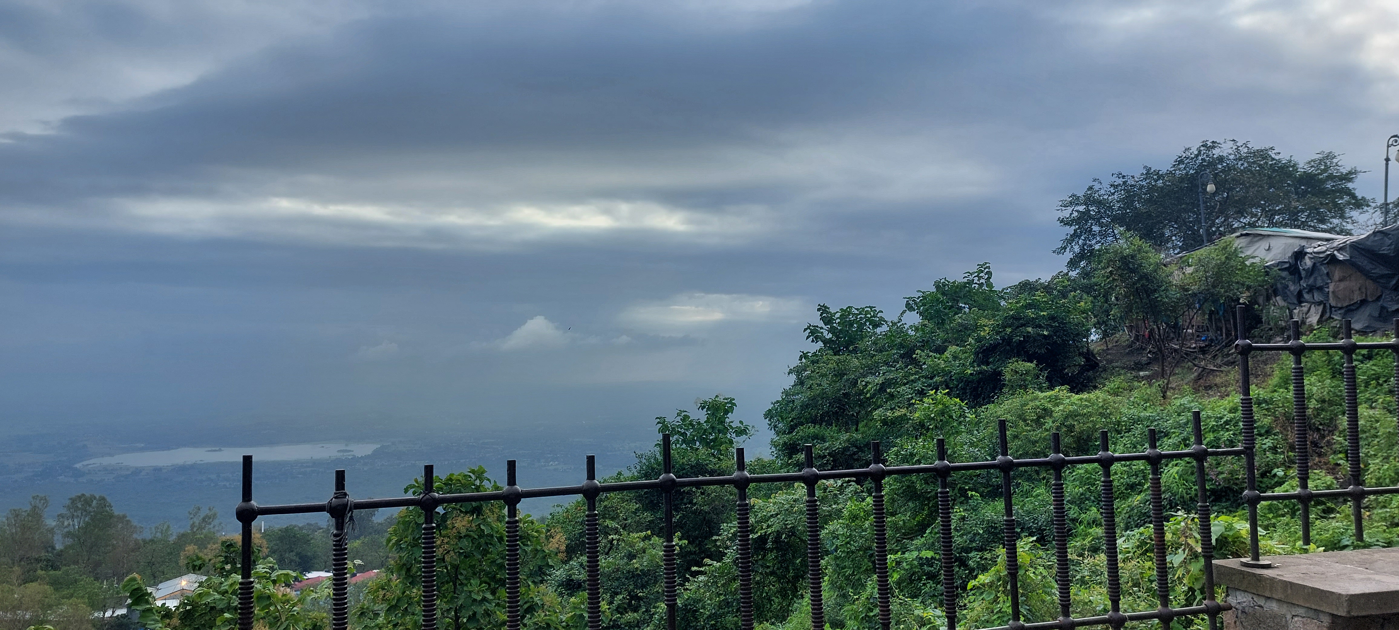 A scenic view of a cloudy sky above a green hillside, with dense trees in the foreground and a distant lake visible through the mist, partially framed by a black metal fence.