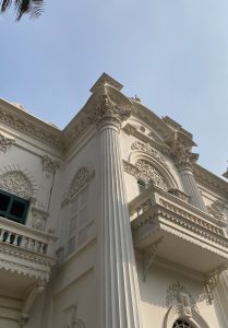 Ornate white building with intricate carvings and tall columns, set against a clear blue sky. The architecture appears grand and historic.