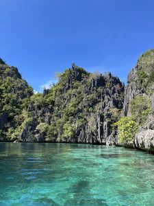 Clear blue-green water in front of tall dark cliffs covered with green trees, under a bright, cloudless blue sky.

