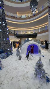 A winter-themed mall decoration featuring an igloo with Santa inside, a penguin figure, artificial snow, and blue-and-white decorated Christmas trees, creating a festive, icy wonderland.
