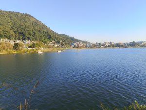 A serene lakeside view featuring calm water reflecting the blue sky, surrounded by lush green hills on one side.