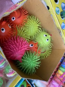 Colorful rubber puffer toys with googly eyes and spiky textures fill a cardboard box. The scene is playful, with red, green, and pink hues.