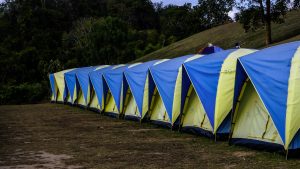 A line of identical blue and yellow dome tents set up outdoors in a grassy field with trees and a hill in the background, suggesting a campsite or organized camping event.