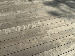 A close-up view of a wooden deck, featuring long, evenly spaced planks secured with small metal screws. The wood shows natural grain patterns and subtle weathering, giving it a rustic, sun-washed look. Soft shadows fall across the boards, adding depth and texture to the simple, minimalistic scene.