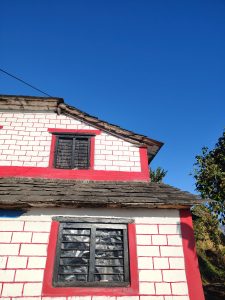 A white house with red accents, black-framed windows with metal grilles, and a wooden shingle roof stands against a clear blue sky.