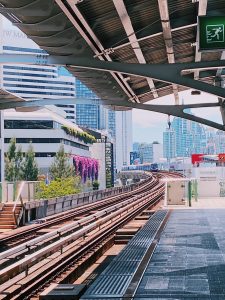A photo from a raised train station platform, showing parallel train tracks curving to the right, with a modern urban cityscape, including tall buildings and the 'JW Marriott' sign, visible in the background under an overcast sky.