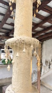 A close-up view of a decorative pillar made of a textured, light brown material, featuring a series of small, diamond-shaped ornaments hanging from thin strings