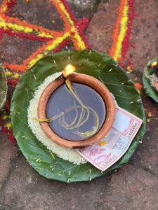 A clay oil lamp sits on a leaf plate with rice and Nepali currency placed beside it during a colorful ritual setup.