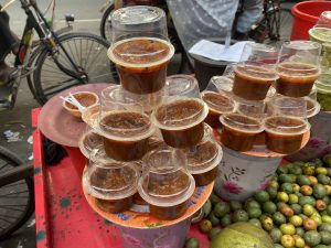 Plastic cups filled with a dark brown sauce are stacked in a pyramid on a street vendor's table. Ripe oranges and limes are scattered beside them.