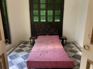 A small room with a single bed, pink blanket, wooden headboard, two side tables, checkered floor, and green-tinted window.