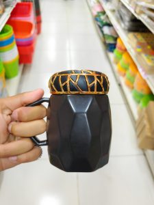 A person's hand is holding a black, faceted mug with a handle, topped with a decorative gold and black ring. 