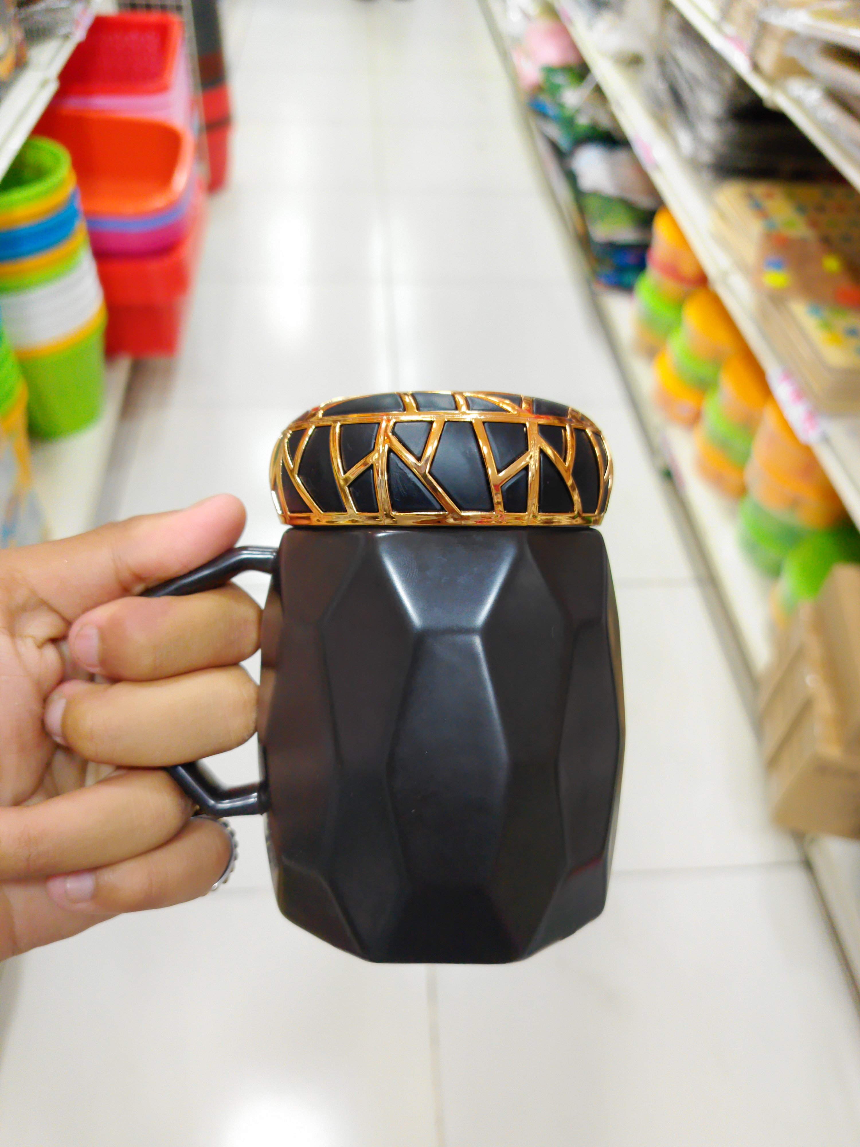 A person's hand is holding a black, faceted mug with a handle, topped with a decorative gold and black ring. 